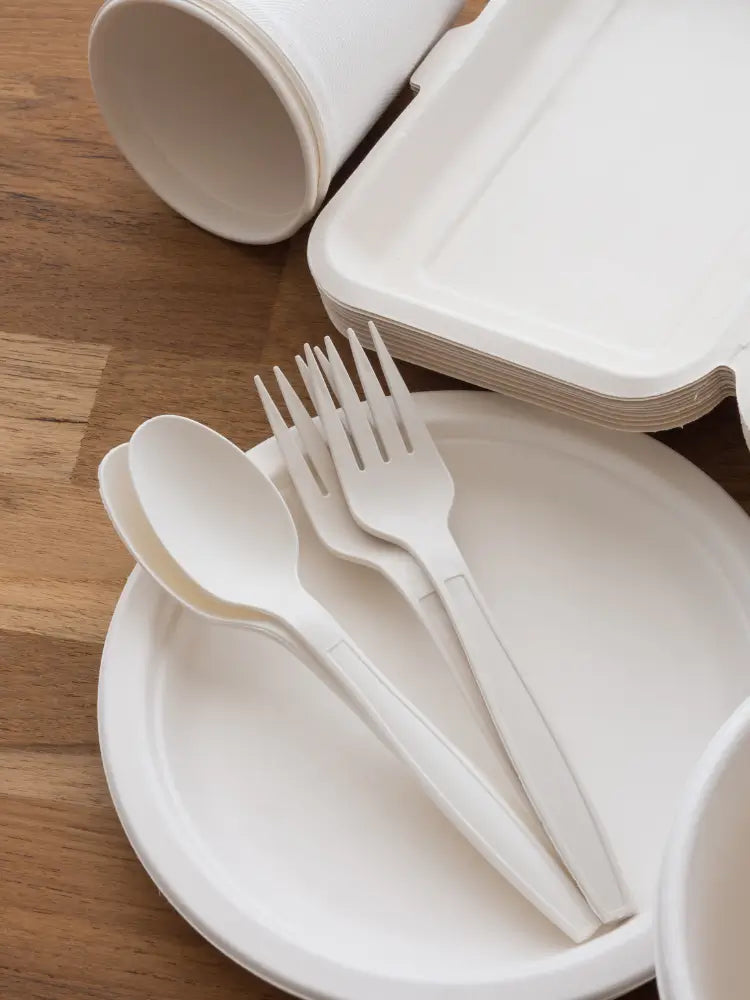 A collection of bagasse plates and bagasse cutlery against wooden countertop.
