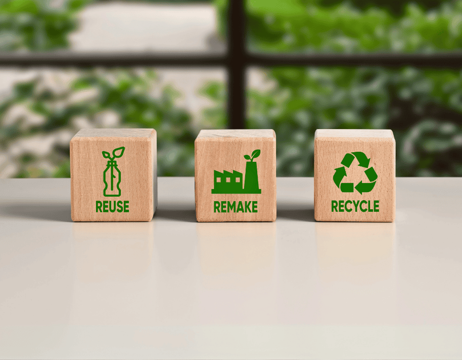 Three wooden blocks with icons and text: REUSE, REMAKE, RECYCLE on a table, greenery in the background.