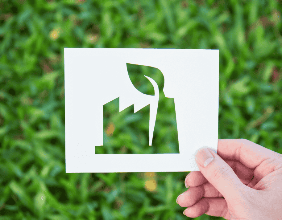 Hand holding a cut-out paper revealing a factory shape against a green grass background.