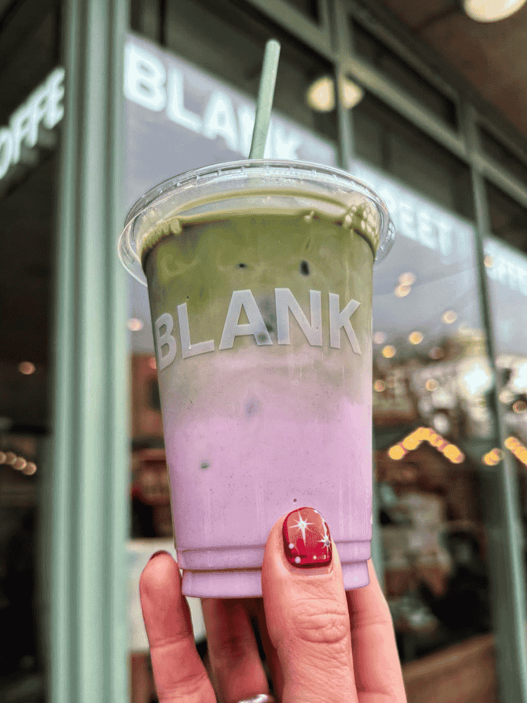A hand holds an iced drink in a clear cold cup. The drink has two layers: green and purple. A café storefront is visible in the background.