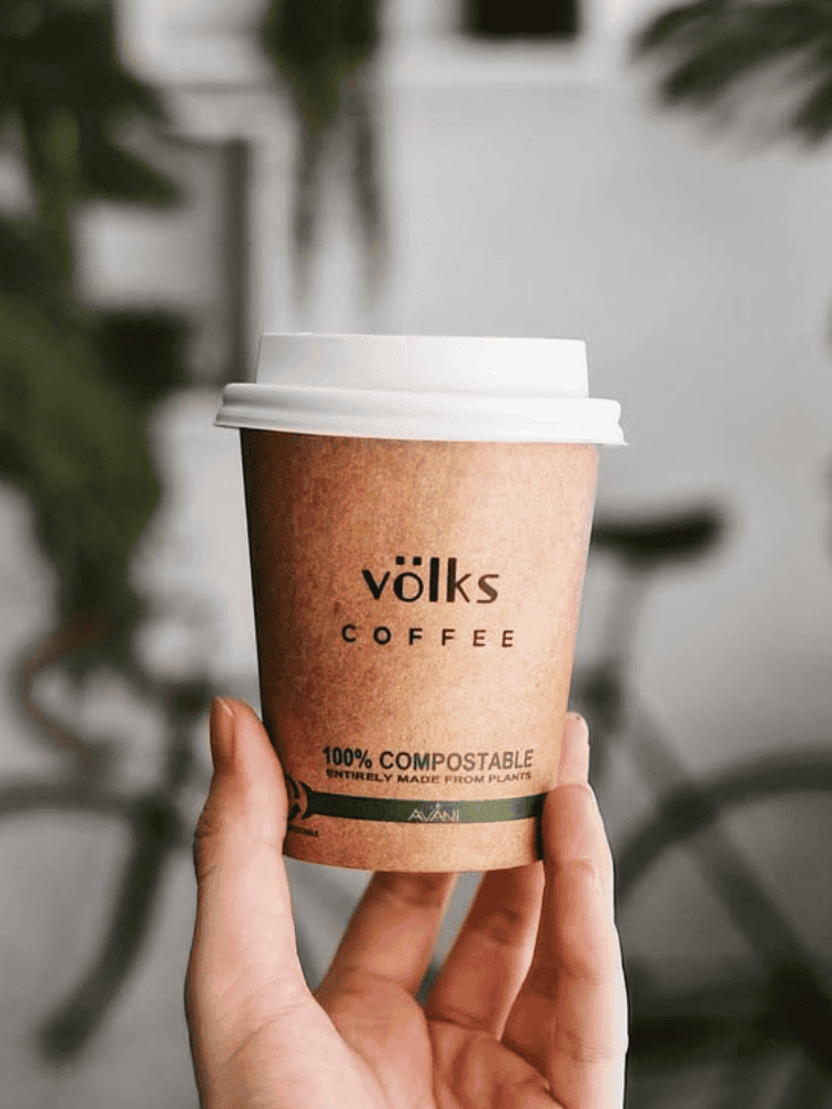 Hand holding a compostable coffee cup labelled Völks Coffee with a blurred bicycle in the background.