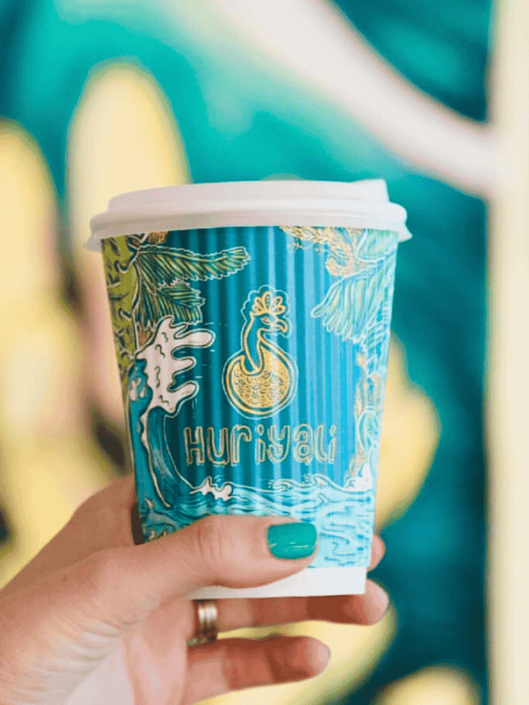 A hand holding a blue paper ripple coffee cup with white lid featuring Huriyali and colourful tropical designs.