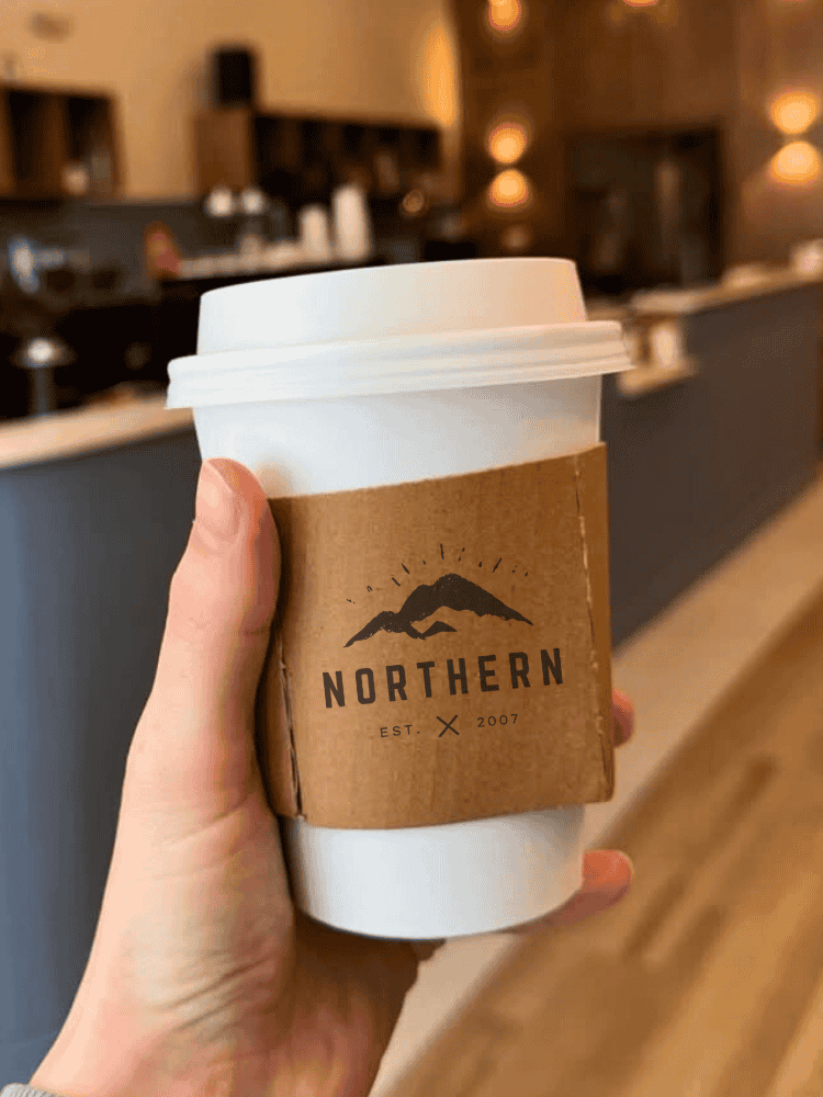 A hand holds a coffee cup with a brown coffee cup sleeve labelled Northern Est. 2007, against the backdrop of a café interior.