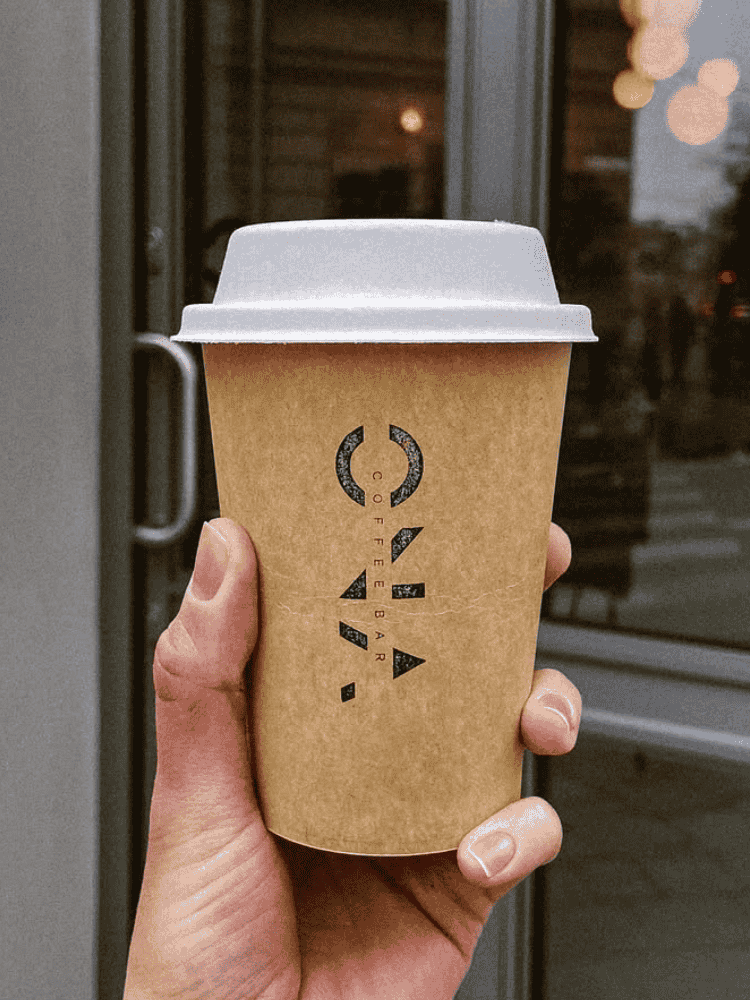 Hand holding a brown double wall coffee cup with a white lid in front of a glass door.