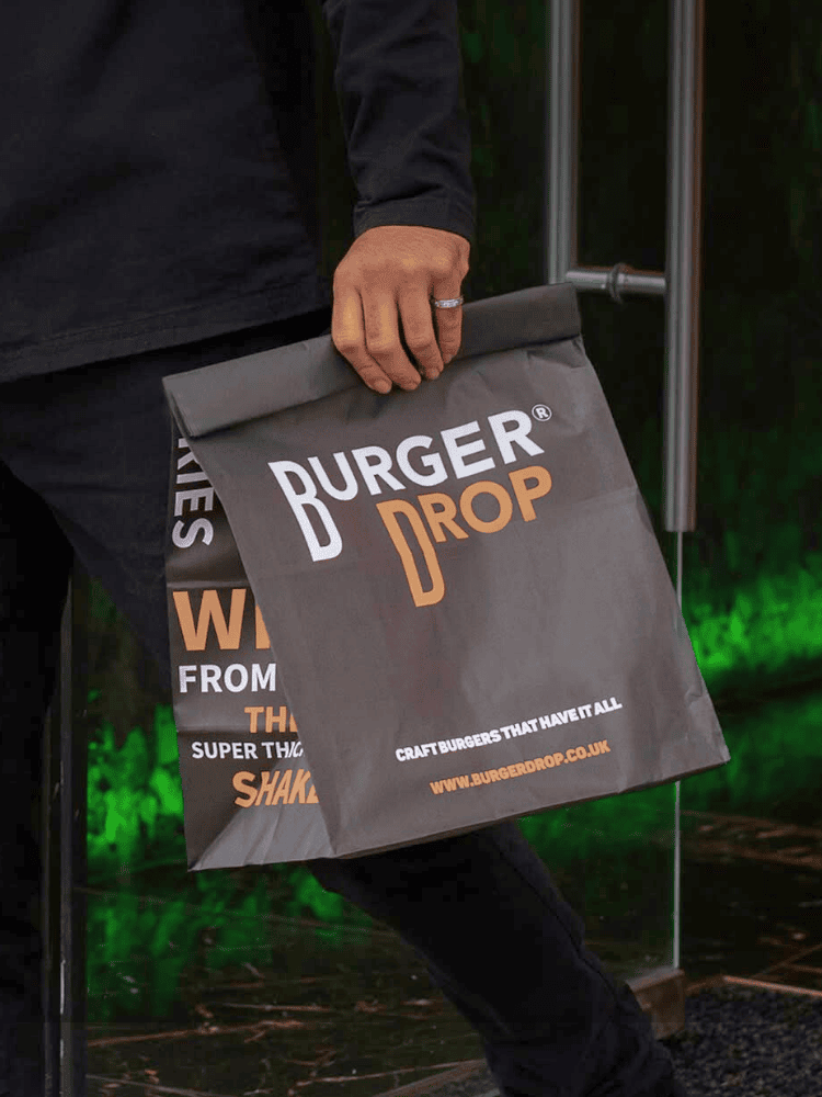 Person holding a black Burger Drop paper takeaway bag, standing near a glass door with a green reflection.