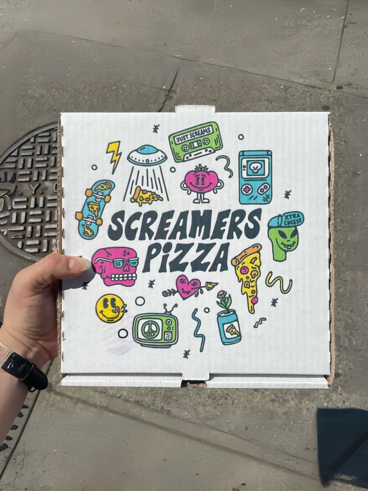 A hand holding a pizza box with colorful illustrations and the text Screamers Pizza on the lid.