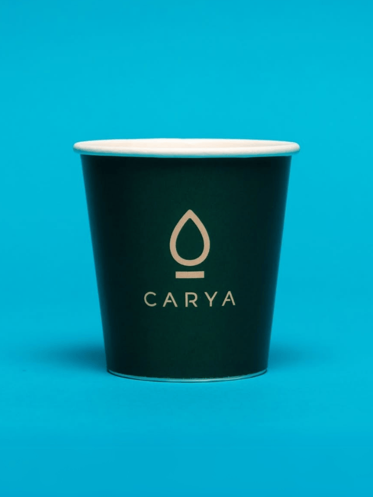 A dark green paper single wall cup with a logo resembling a drop and the word CARYA printed on it, set against a bright blue background.